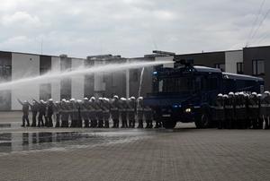 Policjanci Nieetatowych Pododdziałów Prewencji Stojący w tyralierze, po środku stoi armatka wodna, na krańcach szyku stoją policjanci ze strzelbami gładkolufowymi