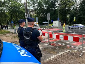 dwóch umundurowanych policjantów stojących przed radiwozem obserwuję otoczenie. W oddali widać policyjny radiowóz. Po lewej stronie w oddali widać patrol straży miejskiej