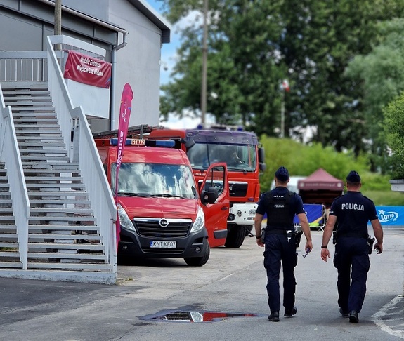 policjanci patrolują teren przed halą lodową  z lewej strony zaparkowane wozy strażackie