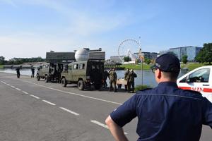 policjant stojący tyłem. Na ulicy stojące samochody wojskowe oraz żołnierze trzymający w rękach skrzynki