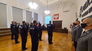 na sali nowi policjanci wypowiadają rotę ślubowania, obok stoją policjanci z kadry kierowniczej, w tle widać wyświetlany slajd z treścią roty oraz sztandar