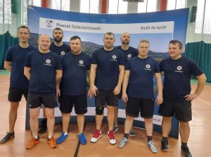 drużyna policjantów biorąca udział w turnieju. Mężczyźni w strojach sportowych  stoją przed banerem na hali sportowej