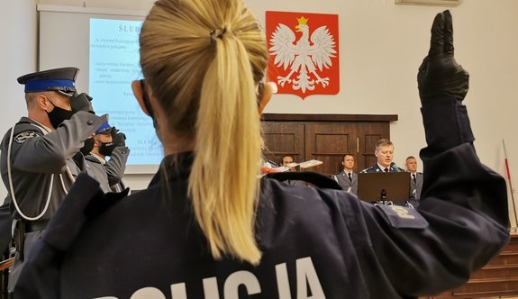 nowo przyjęta policjantka z uniesioną prawą ręką wymawia rotę ślubowania, w tle widać pozostałych uczestników uroczystości