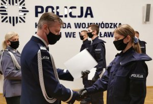 Komendant gratuluje nowym policjantom, wręcza im teczki z aktami mianowania, w tle widać innych policjantów