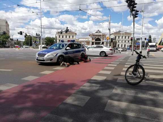 radiowóz blokujący ruch na skrzyżowania, obok ludzie próbujący wydostać kaczkę spod auta,