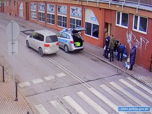 Zdjęcia przedstawiają ulicę, przy której policjanci udzielają poszkodowanej kobiecie pomocy. Na ulicy stoi oznakowany radiowóz