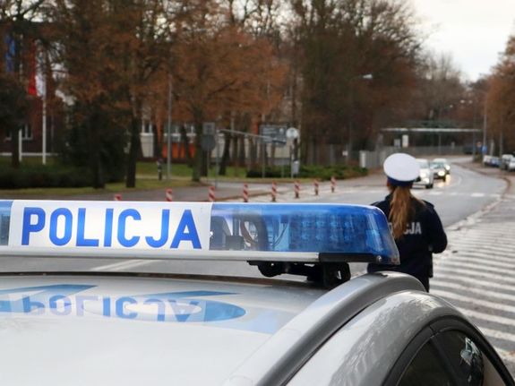 Na dworze przy drodze na pierwszym planie belka od świateł błyskowych z napisem policja, w tle droga  policjantka w białej czapce