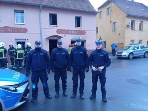 Czterech policjantów stojących na tle budynku. Z tyłu widać radiowozy i strażaków.