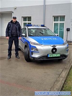 Na zdjęciu policjant Łukasz Warchoł stojący przy radiowozie.