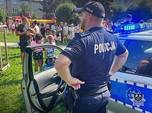 Policjant stojący przy radiowozie, w tle bawiące się podczas festynu dzieci.