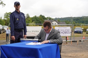 Mężczyzna siedzący na krześle przy stole podpisuje akt erekcyjny. Za nim stoi policjant.