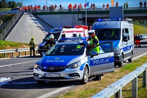 Policjant przy radiowozach w tle sceny z akcji ratunkowej po wypadku - sceny z ćwiczenia służb ratowniczych na drodze S-3 z udziałem dolnośląskich policjantów.