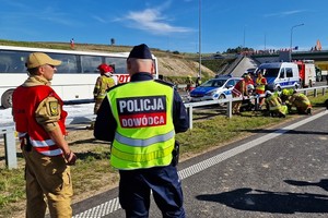 Strażak rozmawia z dowódcą policji w tle strażacy udzielają pierwszej pomocy poszkodowanym - sceny z ćwiczenia służb ratowniczych na drodze S-3 z udziałem dolnośląskich policjantów.