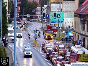 Widok z kamery miejskiej na zatłoczoną ulicę i policjanta kierującego ruchem i strażaków gaszących płonący samochód