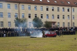 Grupa osób stojących w rzędzie na wzniesieniu przed murawą, a przed nimi samochody, dym i policjanci