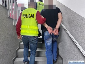 Policjant w kamizelce odblaskowej prowadzi zatryzmanego mężczyznę skutego w kajdanki