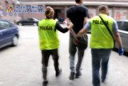 Kadr z filmu, który przedstawia parę policjantów w kamizelkach odblaskowych prowadzących dziedzińcem komendy zatrzymanego mężczyznę.