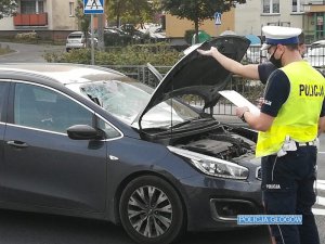 Na zdjęciu widać samochód, który brał udział w wypadku drogowym, ma rozbitą szybę. Policjant widoczny zna zdjęciu wykonuje oględziny pojazdu notując informacje.