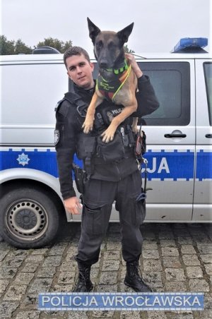 Na zdjęciu policjant z psem służbowym na jego plecach. Z tyłu znajduje się radiowóz.