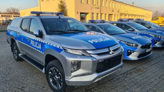 Na zdjęciu 4 oznakowane nowe radiowozy zaparkowane w szeregu. Na pierwszym planie widać samochód marki Mitsubishi L200 koloru srebrno niebieskiego.