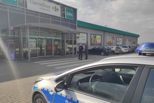 Na zdjęciu widać policjantkę znajdującą się na parkingu marketu, kontrolującą przechodnia. Widać fragment radiowozu oraz zaparkowane samochody.
