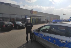 na zdjęciu widać policjantkę, która stoi obok radiowozu na parkingu przed marketem. W tle widać zaparkowane samochody.