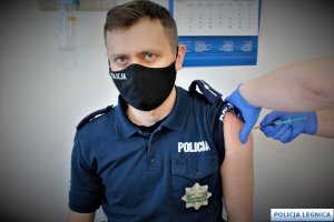 Na zdjęciu widać policjanta siedzącego na fotelu, stojąca obok pielęgniarka podaje szczepionkę