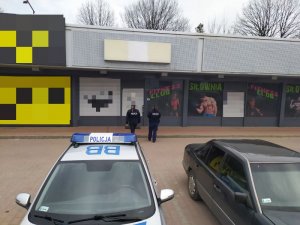 Na zdjęciu widać policjantów wchodzących do lokalu usługowego widać radiowóz i  inne samochody stojące na parkingu