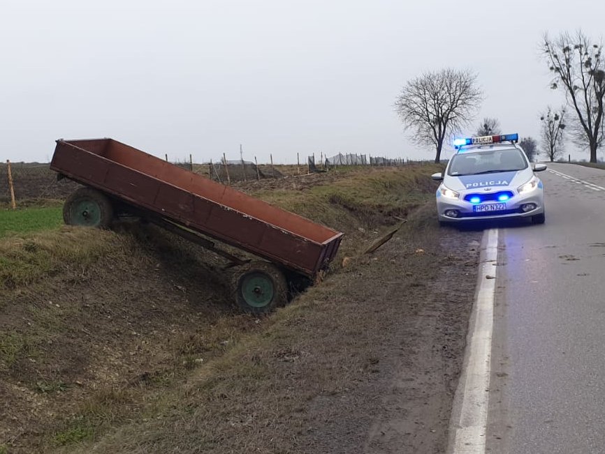 Przyczepa ciągnika w rowie obok policyjny radiowóz z włączonymi lampami błyskowymi.