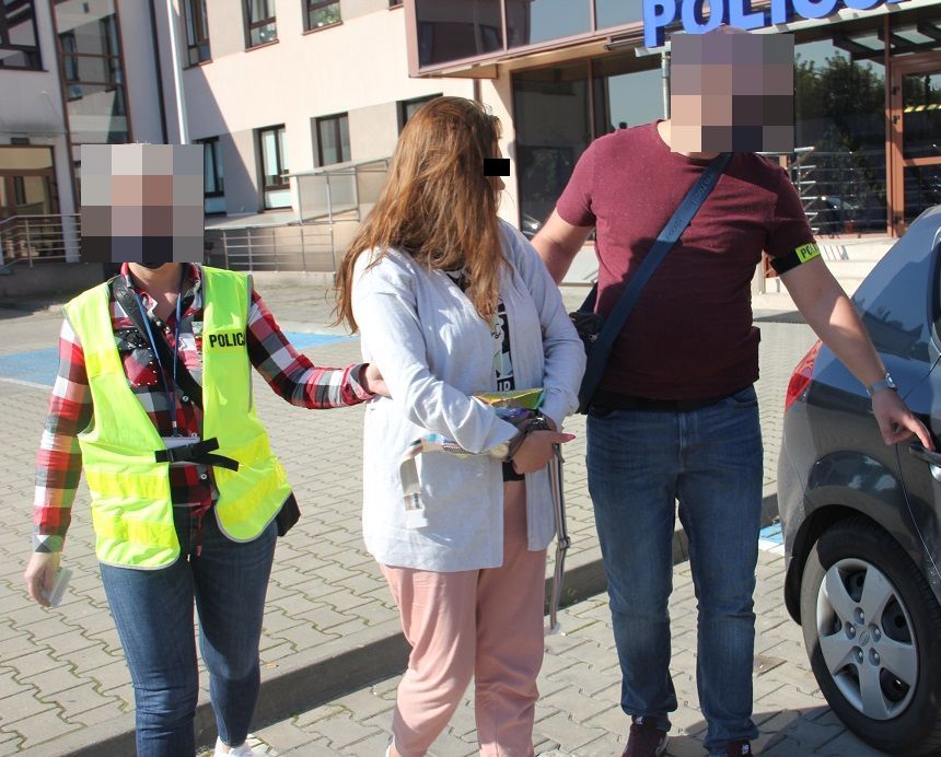 Zatrzymaną kobietę prowadzą do radiowozu policjanci po cywilnemu ubrani z emblematami z napisem policja.  