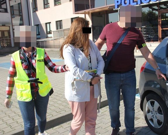 Zatrzymaną kobietę prowadzą do radiowozu policjanci po cywilnemu ubrani z emblematami z napisem policja.