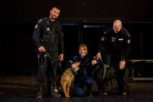 na scenie dwóch stojących policjantów w czarnym mundurze, trzymający dwa psy:jasny i czarny. Pomiędzy nimi klęcząca policjantka w granatowym mundurze