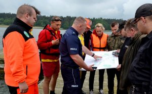 Policjant stoi na pomoście, czyta mapę, a wokół niego stoją inni uczestnicy szkolenia.