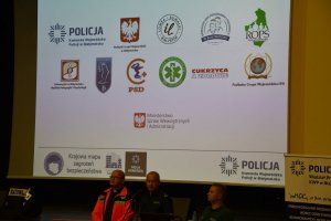 Policjant, ksiądz i ratownik prezentują slajdy promujące bezpieczeństwo osób starszych.