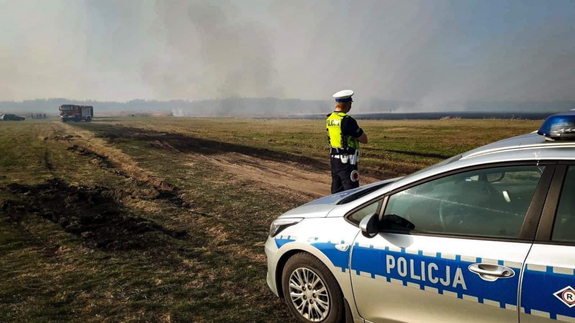 Policjant z radiowozem. W tle palące się łąki.