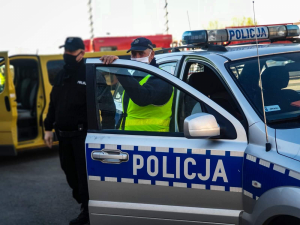 Policjant stoi przy radiowozie. W tle strażacy.