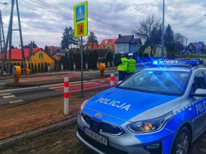 Policjanci stoją przy przejściu dla pieszych na tle radiowozu.