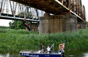 Przewodnik z psem służbowym wisi na linie na moście. Na wodzie jest policyjna łódka z dwoma policjantami i strażakiem.