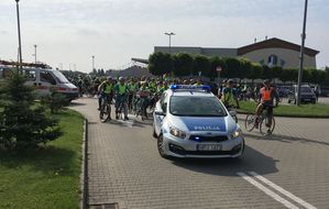 Oznakowany radiowóz z włączonymi sygnałami świetlnymi sto na parkingu przy basenie oleska laguna. Za nim grupa rowerzystów.