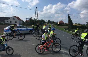 Policjant w kamizelce odblaskowej z zatrzymuje ruch na ul. Opolskiej w Łowoszowie. Przed nim rowerzyści przechodzą i przejeżdżają przez drogę.