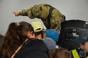 na pierwszym planie głowy dzieci patrzą na opolskiego kontrterrorystę, który pokazuje na coś palcem